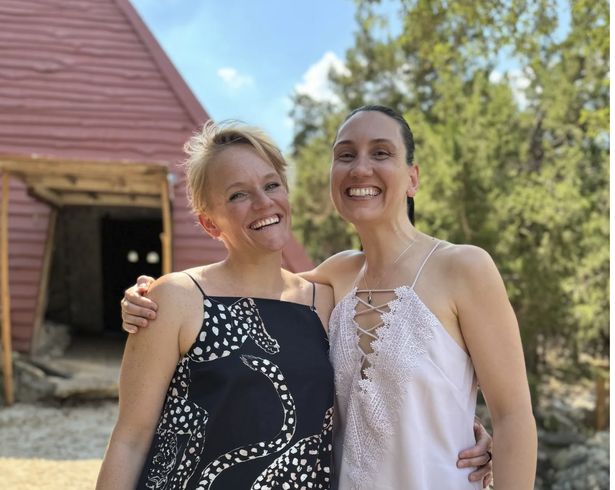 Two women smiling and posing arm-in-arm outdoors on a sunny day, with a red wooden building and trees in the background.