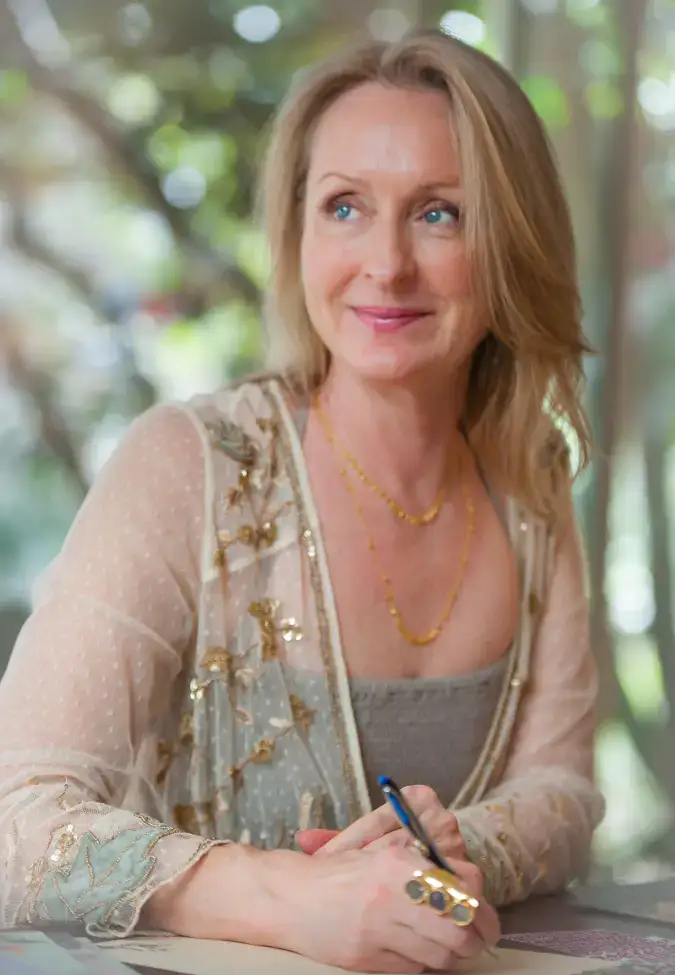 Smiling woman with blonde hair writes with a pen at a table, papers in front, wearing a sheer embroidered blouse and gold jewelry.