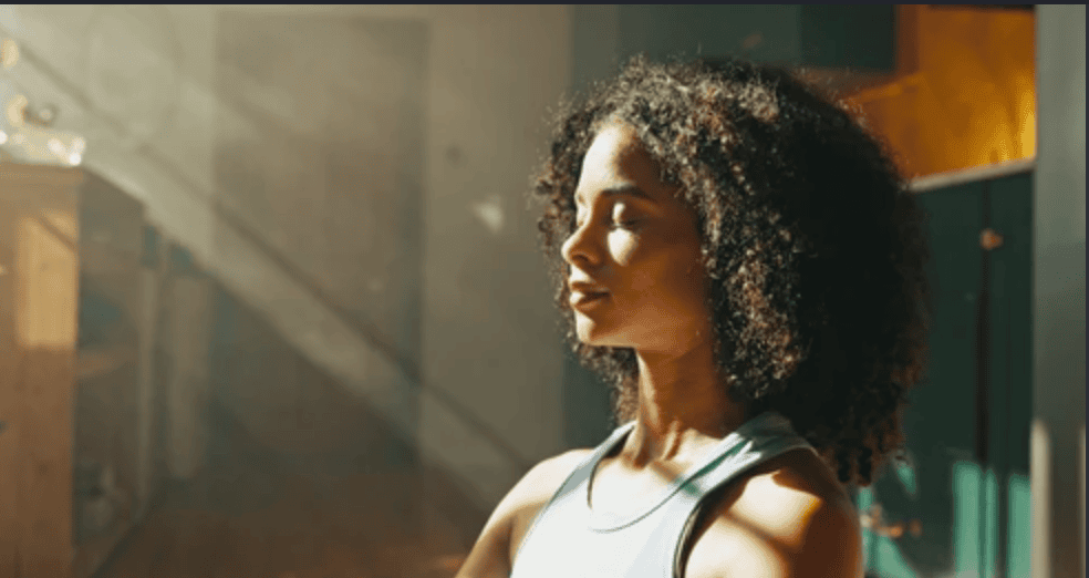 Woman with curly hair wearing a light tank top stands with eyes closed as sunlight shines on her in a bright indoor space.