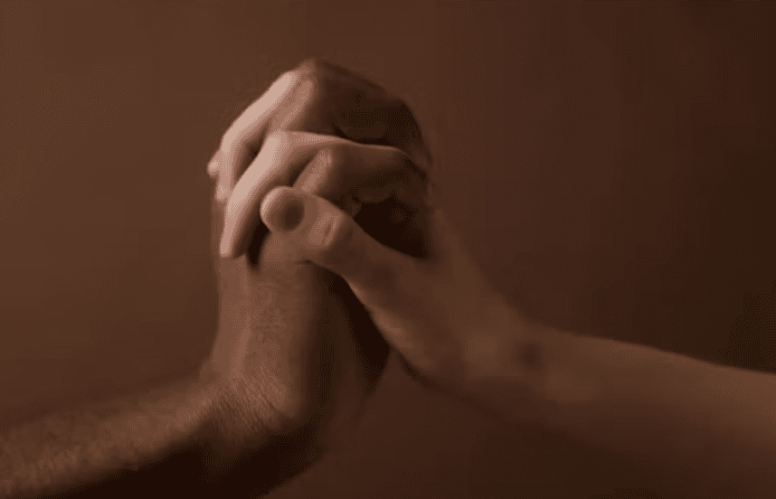 Two hands clasped together in a handshake, one dark-skinned and one light-skinned, against a brown background.
