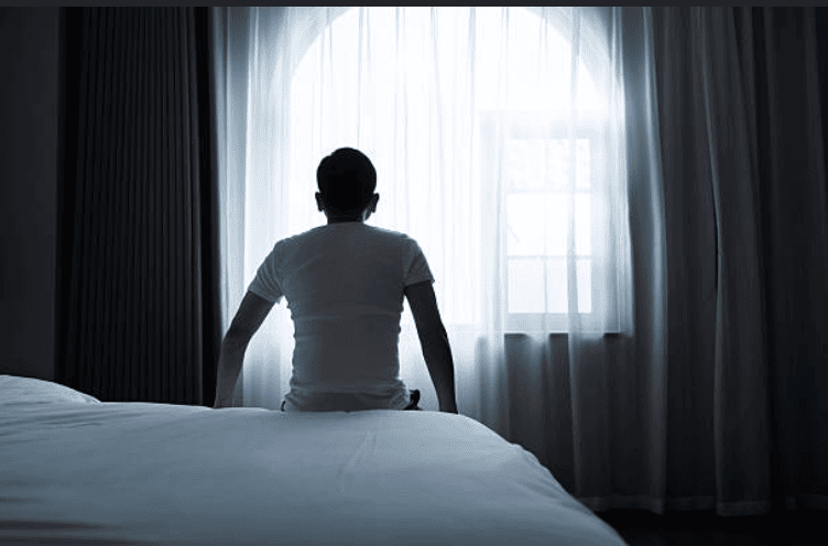 Person sits on a bed with their back to the camera, facing a bright window in a dim bedroom.